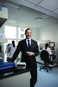 A man in a black suit stands in a lab while holding a piece of equipment. Two women in white lab coats sit at computers in the lab behind him.